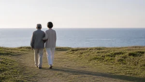 海の見える芝生の道を、腕を支えられてゆっくり歩く年配の人の後ろ姿