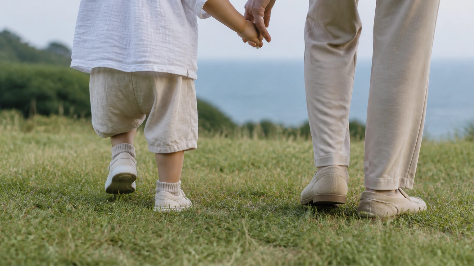 芝生の上で手をつないで歩く、小さな子どもと大人の足元